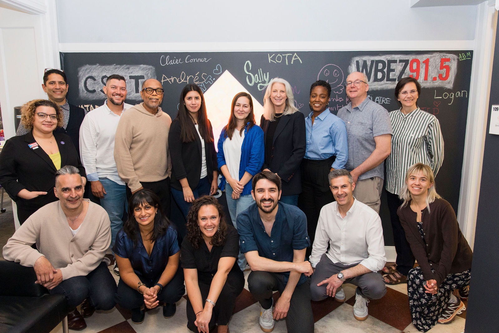A group photo of 16 people posed in two rows in front of a black chalkboard. The front row shows six people sitting or kneeling, while the back row shows ten people standing. The chalkboard behind them contains various handwritten names and text in chalk. The group is standing in the entryway to Knight Lab, with people wearing a mix of business casual attire in various colors including blue, black, white, and striped patterns.