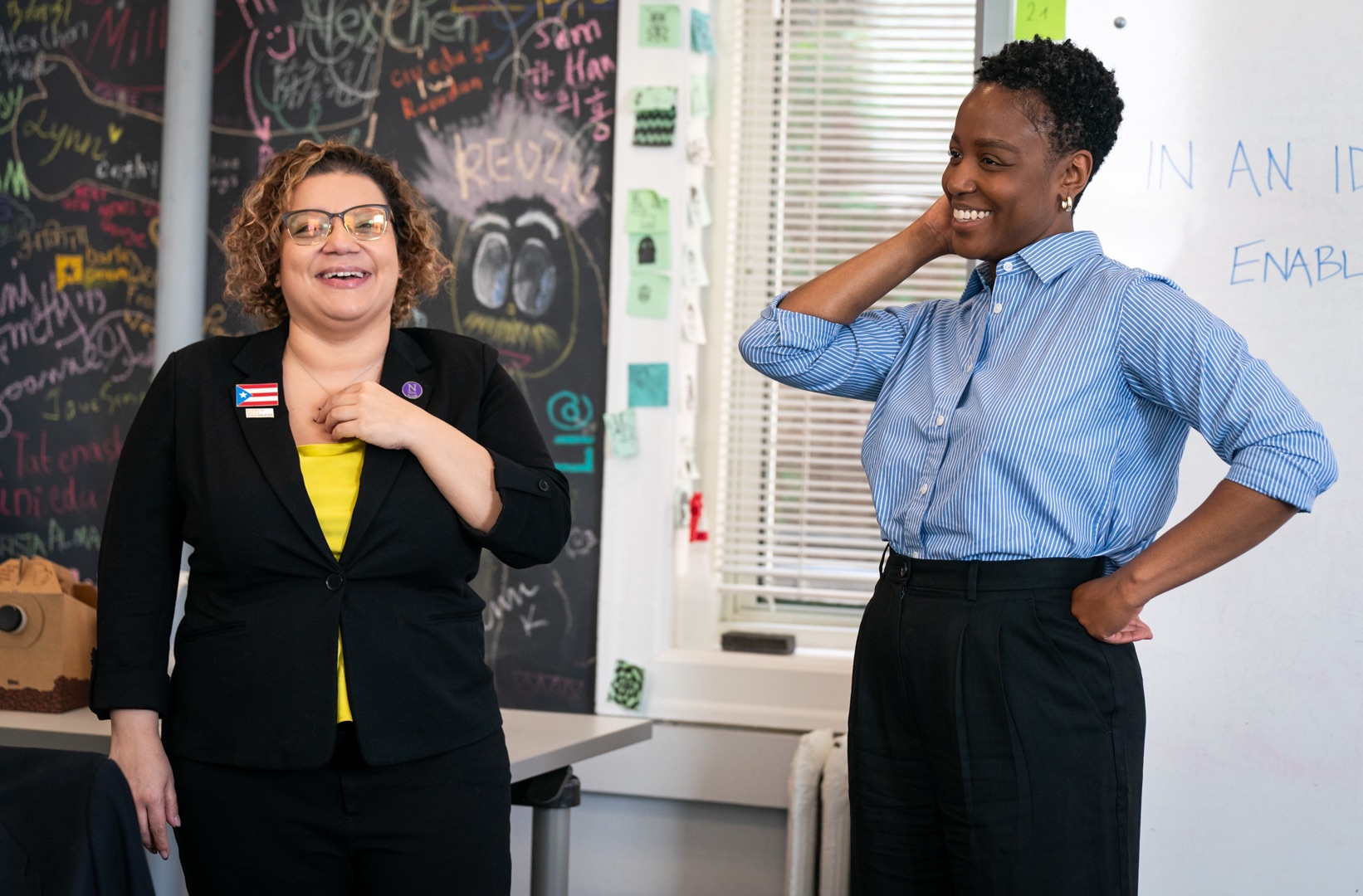 Two people having a conversation in front of a black chalkboard wall covered in colorful chalk writing and drawings. The person on the left wears a light blue button-down shirt and is smiling, while the person on the right wears a white collared shirt and appears to be speaking with hands gesturing. The chalkboard behind them shows the Knight Lab diamond logo, labeled, along with many names of visitors written all over the board, including on top of the logo.