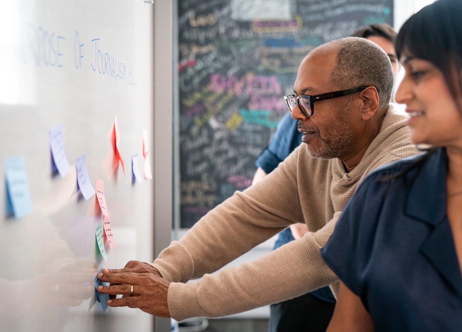 A person wearing glasses and a beige sweater places colorful sticky notes on a whiteboard while another person in a dark blue shirt looks on. The whiteboard has ‘PURPOSE OF JOURNALISM’ written at the top in blue text, with various colored sticky notes arranged below. In the background, there's a black chalkboard covered with colorful chalk writing and drawings.