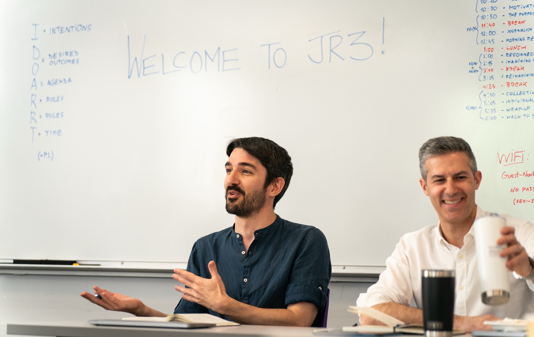 Two men sitting at a conference table in a classroom setting, with one man in a dark blue collared shirt gesturing with his hands while speaking, and another man in a white shirt smiling beside him. A whiteboard in the background shows ‘WELCOME TO JR3!’ and various handwritten notes including schedules and agenda items.